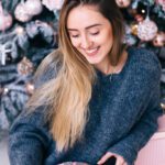 Cute Girl Sitting Against The Christmas Tree And Holding The Box