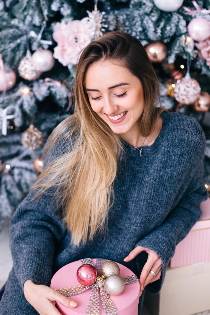 Cute Girl Sitting Against The Christmas Tree And Holding The Box